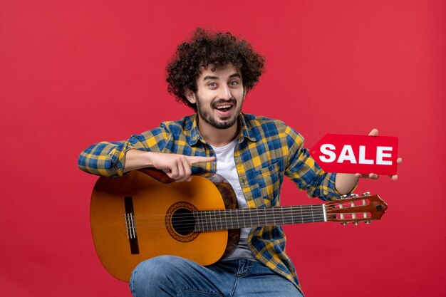 Front view young male sitting with guitar on red wall play music performance musician color applause live concert sale