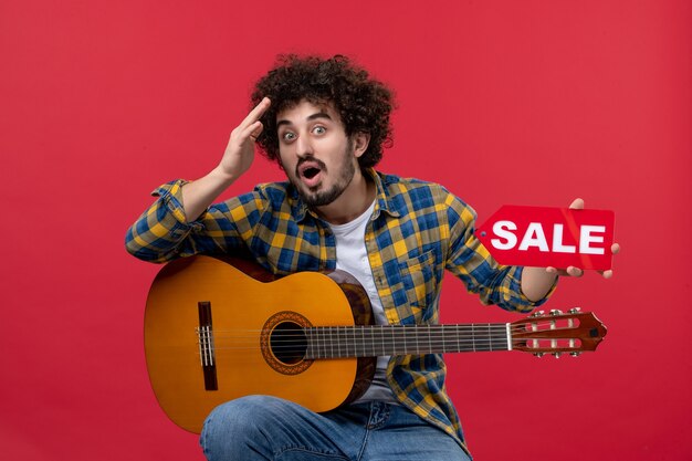 Front view young male sitting with guitar on red wall play music performance musician color live concert sale