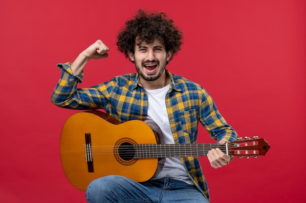 Front view young male sitting with guitar rejoicing on red wall play concert live band music color musician applause