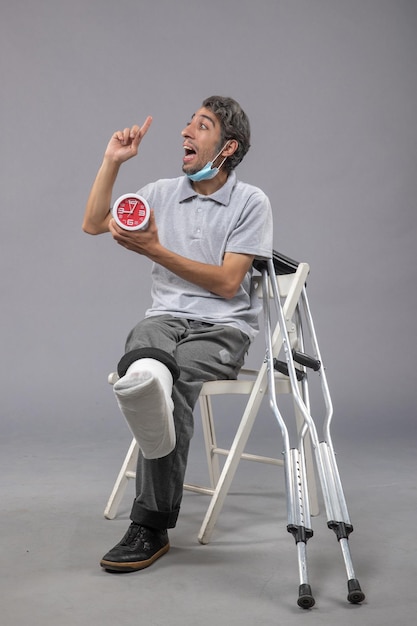 Vista frontale giovane maschio seduto con fasciatura legata a causa di un piede rotto e tenendo l'orologio sul muro grigio torsione gamba dolore al piede lussazione incidente