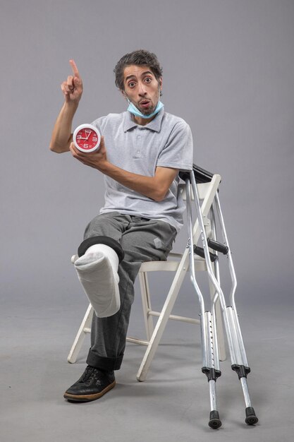 Front view young male sitting with tied bandage due to broken foot and holding clock on a grey wall twist leg foot pain accident male