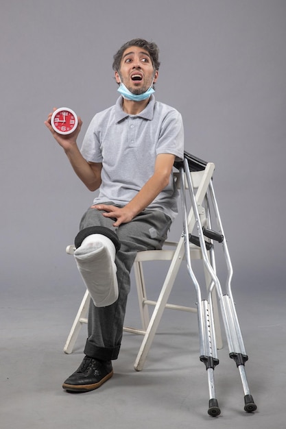 Vista frontale giovane maschio seduto con fasciatura legata a causa di un piede rotto e tenendo l'orologio sul muro grigio torsione dolore piede incidente gamba maschio