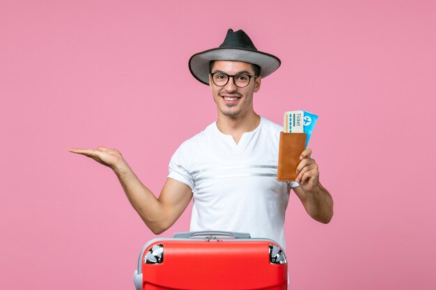Вид спереди молодой мужчина улыбается и держит билеты на самолет на розовом столе, путешествие, поездка, самолет, мужское фото, цвет, отпуск