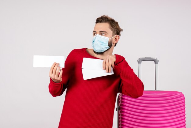Front view young male in sterile mask holding tickets on white wall voyage covid- flight trip color emotion virus