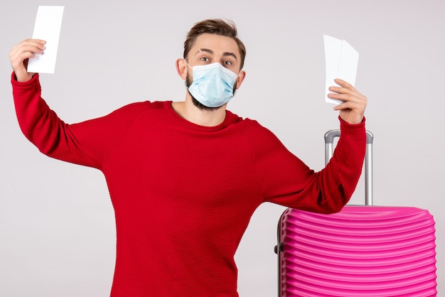 Front view young male in sterile mask holding tickets on white wall voyage covid- flight trip vacation color emotion virus