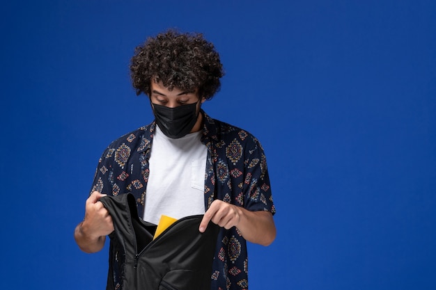 Front view young male student wearing black mask and holding backpack on light blue background.