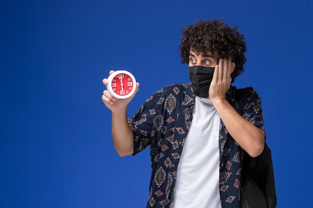 Front view young male student wearing black mask with backpack holding clocks on blue background.