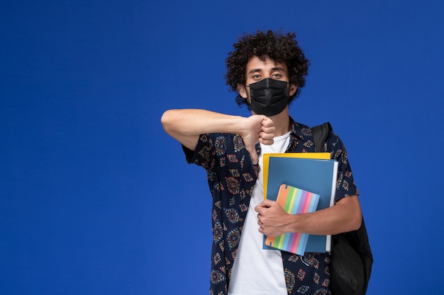 Vista frontale giovane studente maschio che indossa la maschera nera con zaino in possesso di quaderno e file su sfondo blu.