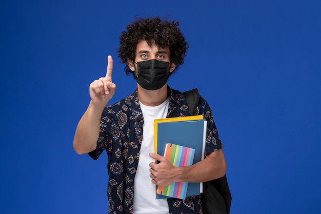 Vista frontale giovane studente maschio che indossa la maschera nera con zaino in possesso di quaderno e file su sfondo blu.