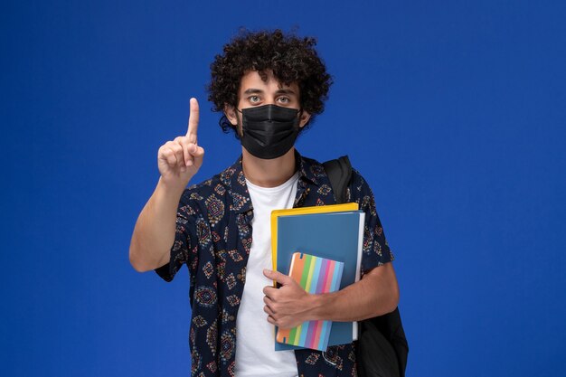 Front view young male student wearing black mask with backpack holding copybook and files on blue background.