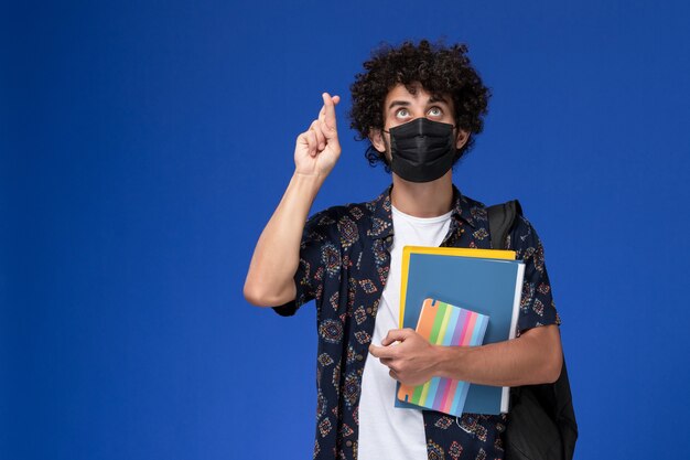 Front view young male student wearing black mask with backpack holding copybook and files on the blue background.