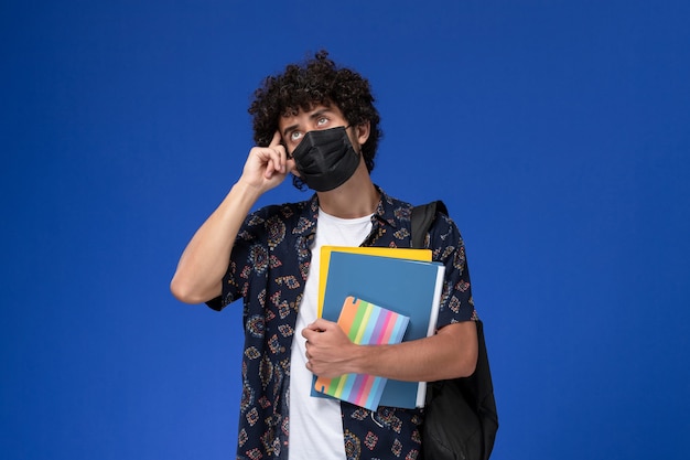 Молодой студент-мужчина вид спереди в черной маске с рюкзаком, держащим файлы и тетрадь с прописями, думая на синем столе.