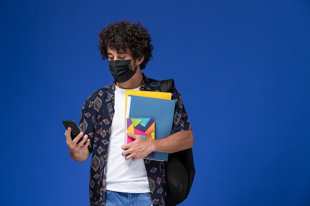 青い背景にファイルと電話を保持しているバックパックと黒いマスクを身に着けている正面図若い男子学生。