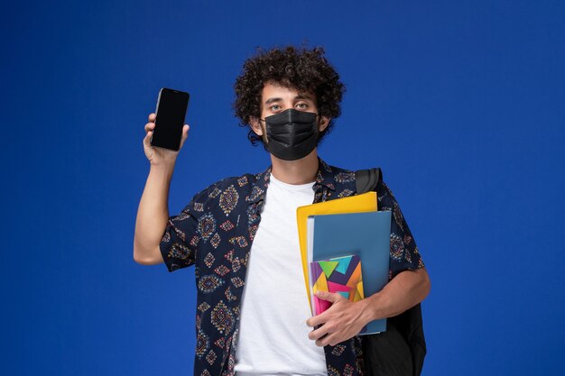 Front view young male student wearing black mask with backpack holding files and phone on the blue background.