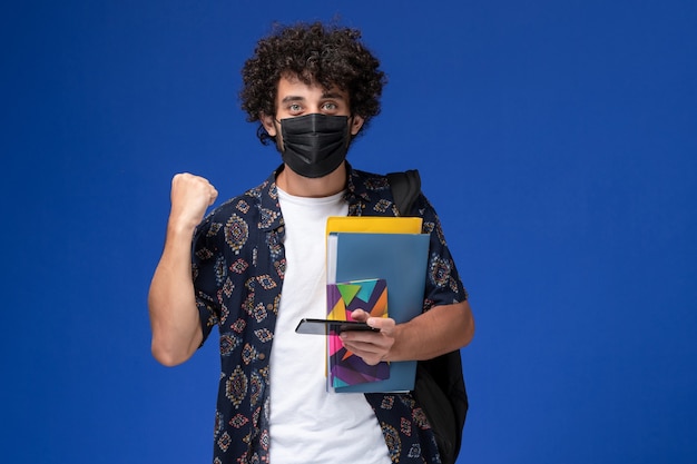 Front view young male student wearing black mask with backpack holding files and using his phone rejoicing on blue background.