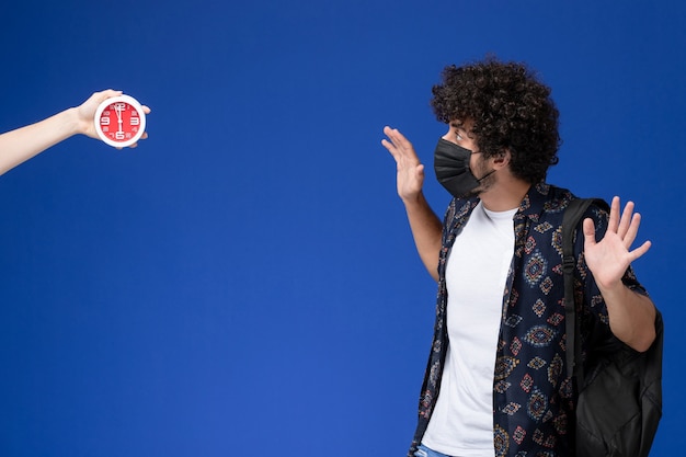 Front view young male student wearing black mask with backpack scared of clocks on light-blue background.