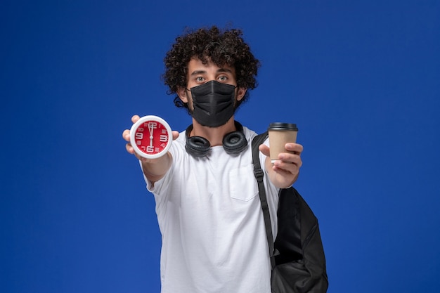 Giovane studente maschio di vista frontale in maglietta bianca che porta maschera nera e che tiene tazza di caffè con l'orologio su fondo azzurro.