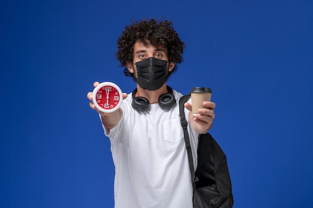 검은 마스크를 착용 하 고 밝은 파란색 배경에 시계와 커피 컵을 들고 흰색 t- 셔츠에 전면보기 젊은 남성 학생.