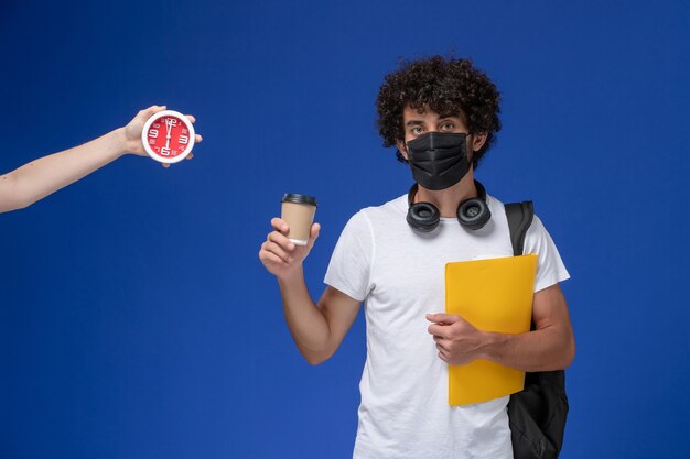 Front view young male student in white t-shirt wearing black mask and holding yellow files and coffee on blue background.