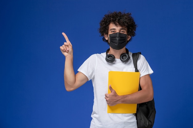 Free photo front view young male student in white t-shirt wearing black mask and holding yellow files on light-blue background.