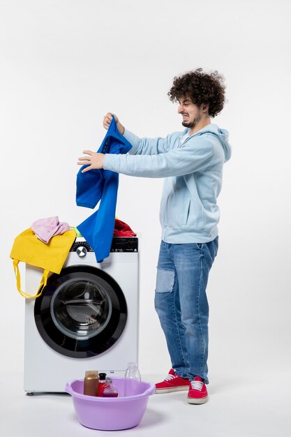 흰 벽에 있는 세탁기에서 깨끗한 옷을 꺼내는 젊은 남성의 전면 모습