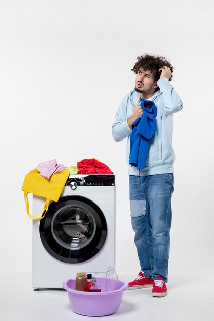 흰 벽에 있는 세탁기에서 깨끗한 옷을 꺼내는 젊은 남성의 전면 모습
