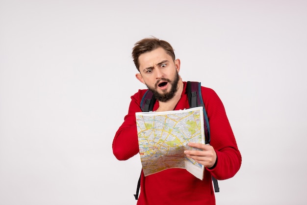 Вид спереди молодой мужчина-турист с рюкзаком, изучающий карту на белой стене, самолет, город, отпуск, эмоция, человеческий цвет, туристический маршрут