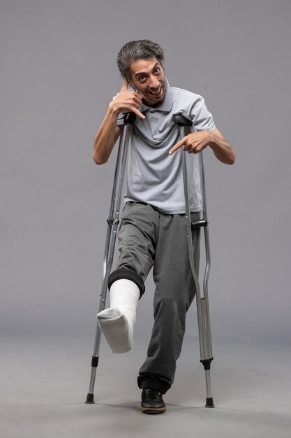 灰色の机の上の足の骨折のために松葉杖を使用している正面図若い男性痛み足が壊れた事故の足を無効にする