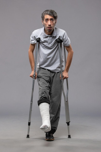Вид спереди молодой мужчина, использующий костыли из-за сломанной ноги на серой стене, боль отключил ногу, сломанную ногу в результате несчастного случая