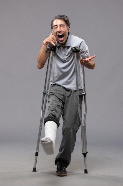 灰色の壁の痛みの足の壊れた足のために松葉杖を使用している正面図の若い男性は事故の足を無効にします