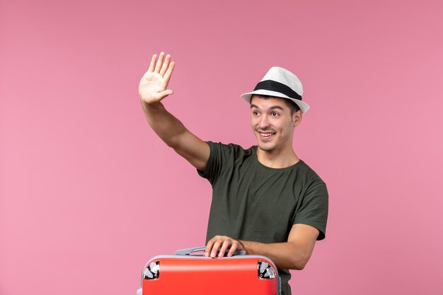 ピンクのスペースに手を振って赤いバッグを運ぶ休暇中の若い男性の正面図