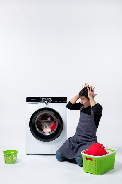 Front view of young male waiting until the end of clothes washing on a white wall