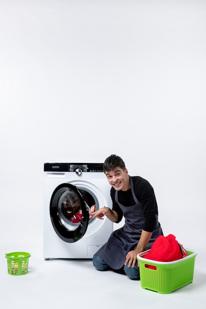 白い壁に洗濯機の助けを借りて服を洗う若い男性の正面図