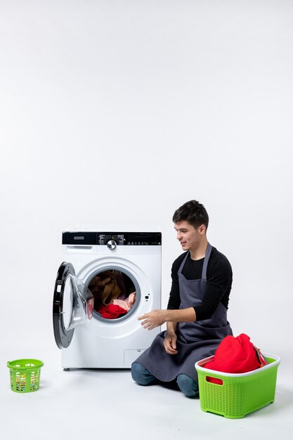 Front view of young male washing clothes with the help of washing machine on white wall