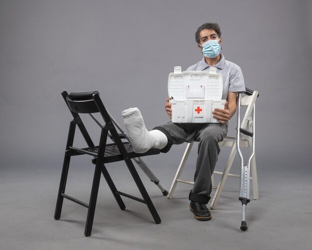 Front view young male wearing mask and sitting with bandaged broken foot on a grey wall twist male broke leg pain foot