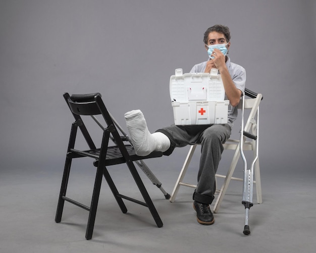 Front view young male wearing mask and sitting with bandaged broken foot on grey wall twist male broke leg pain foot