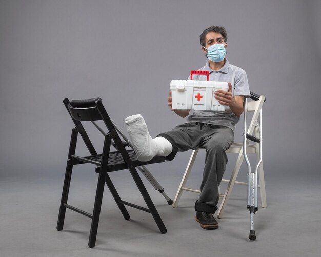 Front view young male wearing mask and sitting with bandaged broken foot on grey wall twist male broke pain foot leg