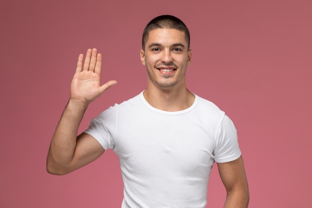 Front view young male in white shirt asmiling and posing with raised hand on the pink background