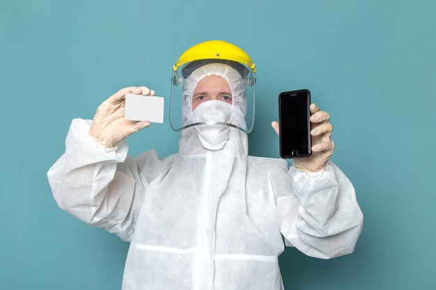 A front view young male in white special suit and yellow special helmet holding card and phone on the blue wall man suit danger special equipment color