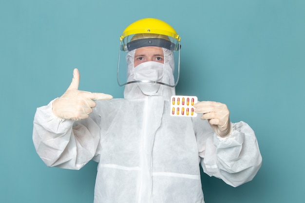 A front view young male in white special suit and yellow special helmet holding pills on the blue wall man suit danger special equipment color