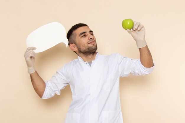 Front view young male in white suit wearing gloves holding white sign and apple on beige