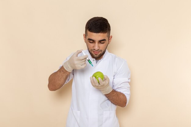 Front view young male in white suit wearing gloves injecting apple on beige