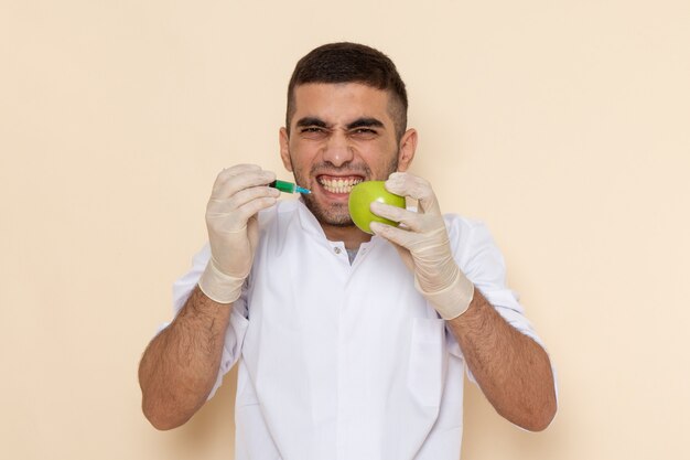 Front view young male in white suit wearing gloves injecting apple on beige