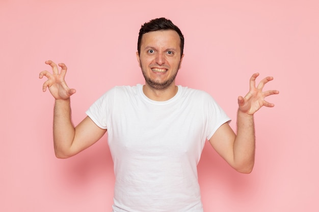A front view young male in white t-shirt and blue jeans posing with funny expression on the pink desk man color emotion pose