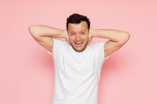 A front view young male in white t-shirt covering his ears on the pink desk man color emotion pose