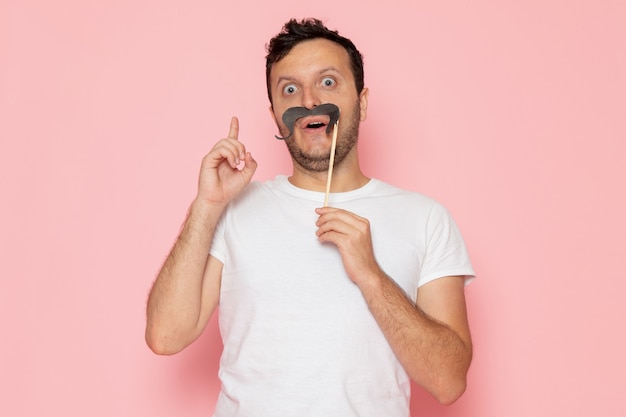 A front view young male in white t-shirt holding fake mustache on the pink desk man color emotion pose