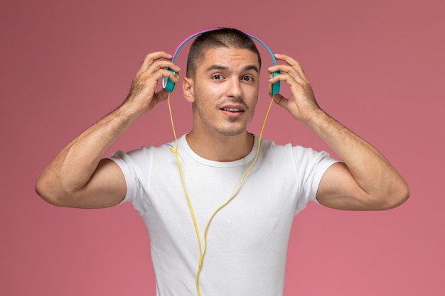 Вид спереди молодой мужчина в белой футболке, слушающий музыку через наушники на розовом фоне