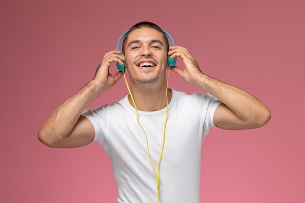 Вид спереди молодой мужчина в белой футболке, слушающий музыку через наушники с улыбкой на розовом фоне
