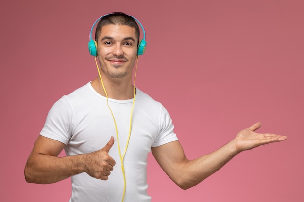 Front view young male in white t-shirt listening to music via his earphones smiling on light-pink background 