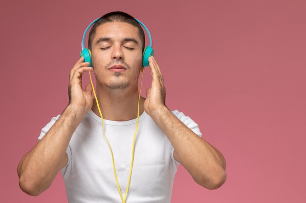 ピンクの背景に彼のヘッドフォンを介して音楽を聴く白いtシャツの正面の若い男性